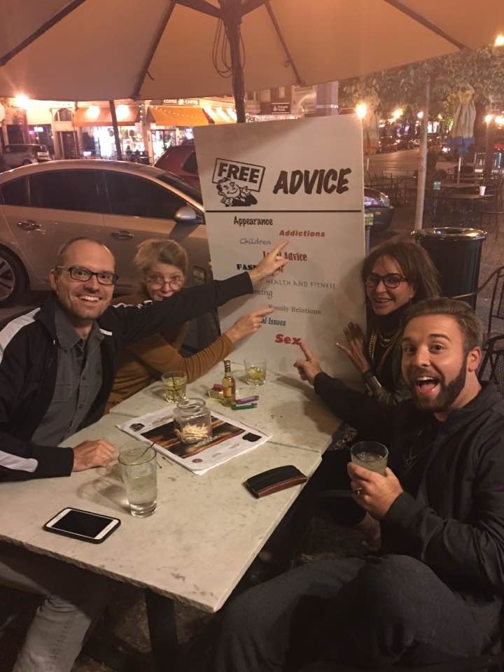 Four visitors smiling and pointing at topics on the Free Advice sign while seated around the outdoor advice table.
