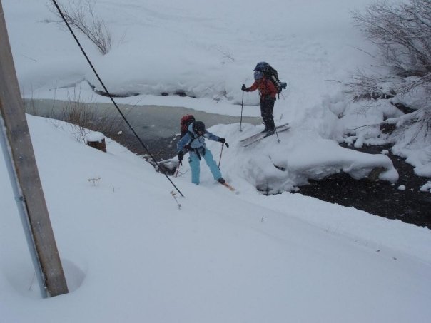 Kippy Hoene climbing out of a snowy creek beside a solid snow bridge while another skier watches.